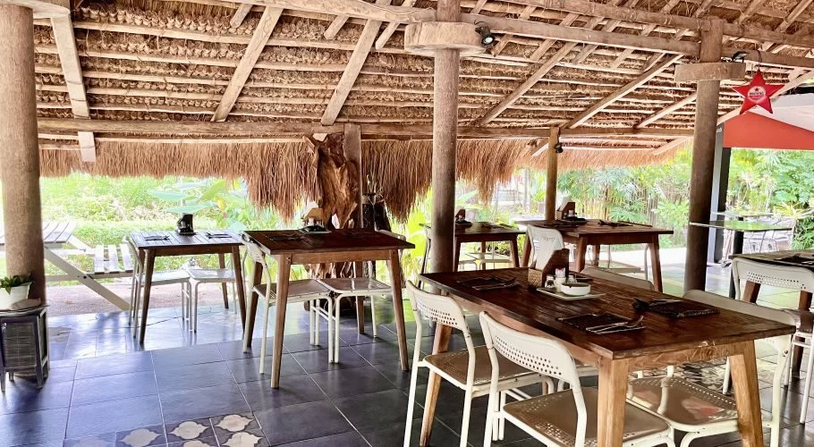 The open-air dining area of Gula Garam Restaurant features wooden tables, white chairs, a dark tiled floor, and a natural thatched roof, with green foliage visible outside.