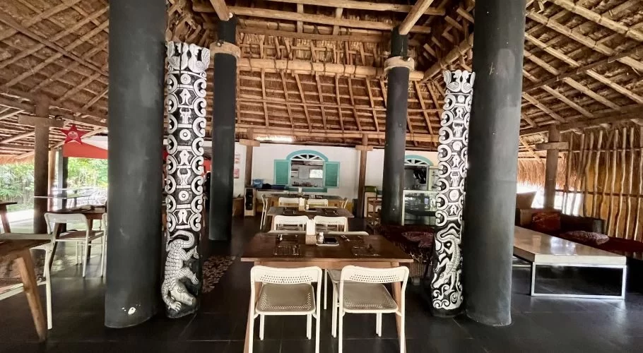 Visible is the interior of a restaurant with a high thatched roof, dark pillars, and wooden tables with white chairs. Some pillars feature white traditional carvings, and green foliage is seen outside the open sides.