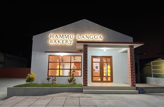 The Hammu Langga Bakery building glows warmly at night, featuring a white facade, illuminated sign, large windows, and a brick-patterned entrance pillar.