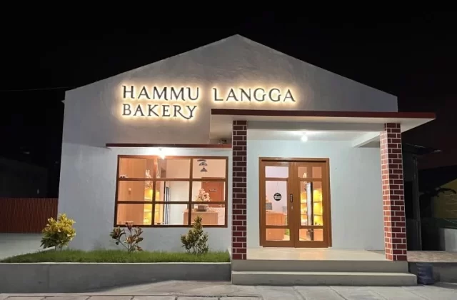 The Hammu Langga Bakery building glows warmly at night, featuring a white facade, illuminated sign, large windows, and a brick-patterned entrance pillar.