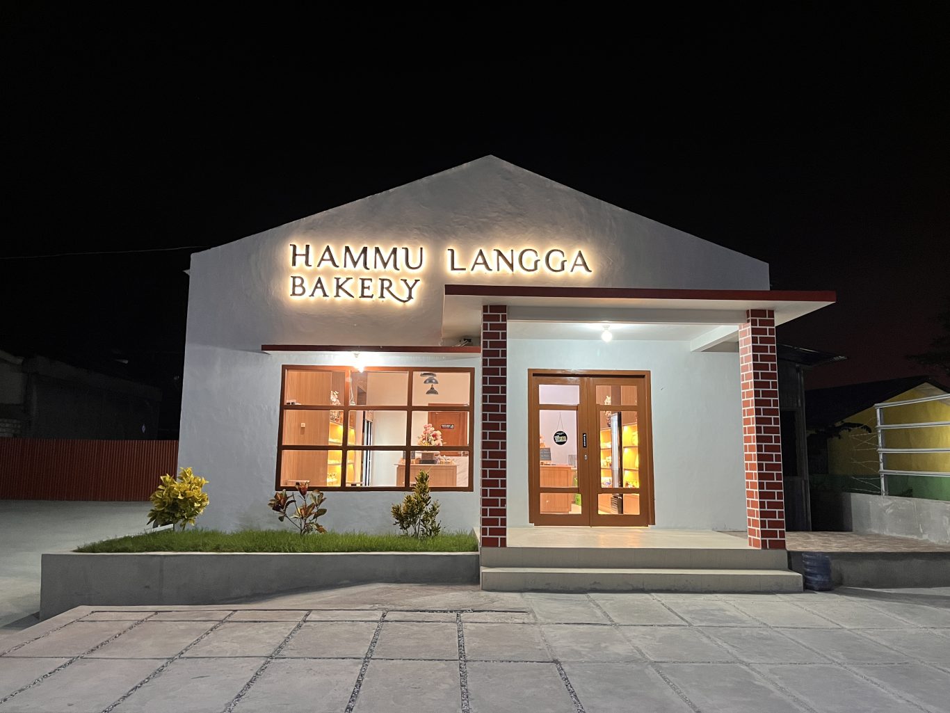 The Hammu Langga Bakery building glows warmly at night, featuring a white facade, illuminated sign, large windows, and a brick-patterned entrance pillar.