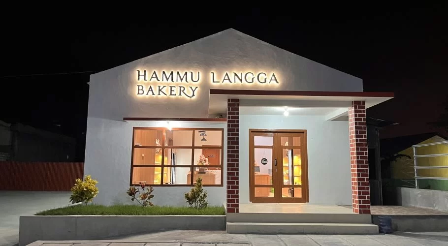 The Hammu Langga Bakery building glows warmly at night, featuring a white facade, illuminated sign, large windows, and a brick-patterned entrance pillar.