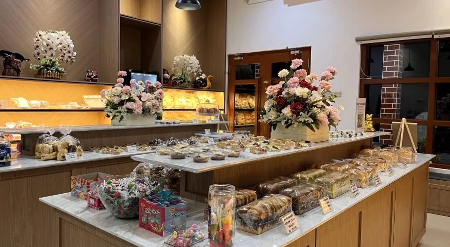 A well-lit bakery interior features multiple display counters filled with various packaged breads, pastries, and sweets, adorned with large floral arrangements.
