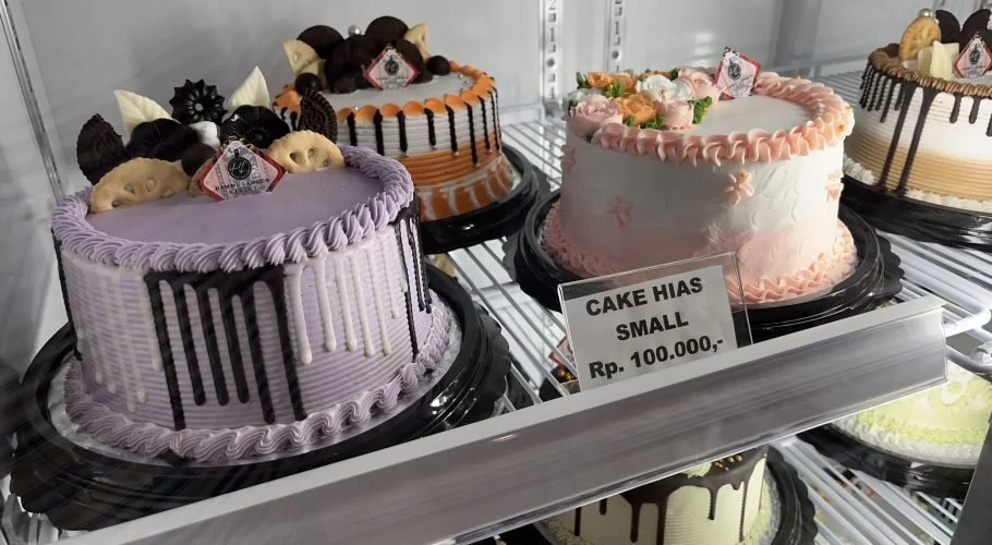 Multiple round, decorated cakes in various colors like purple, pink, and orange are displayed on shelves inside a brightly lit bakery case.