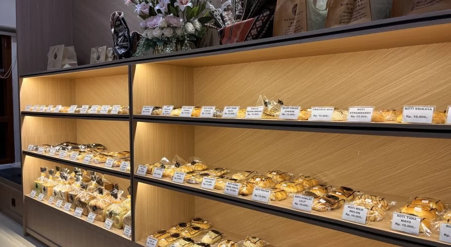Modern wooden bakery shelves display rows of individually wrapped pastries and breads with price tags, illuminated by warm golden lighting.