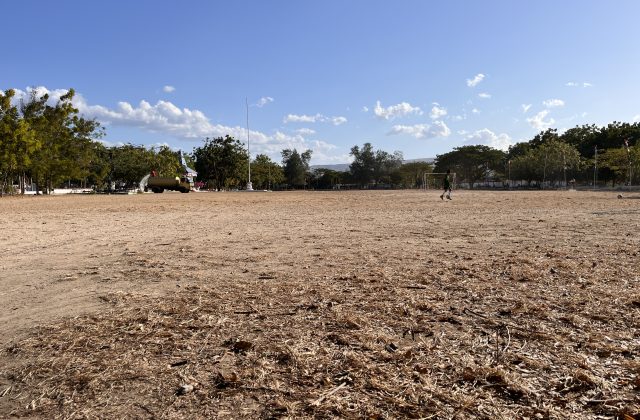 A large, dry, brown field at Heroes Square in Waingapu features a person in a green shirt near a soccer goal, with trees and a flagpole under a blue sky.