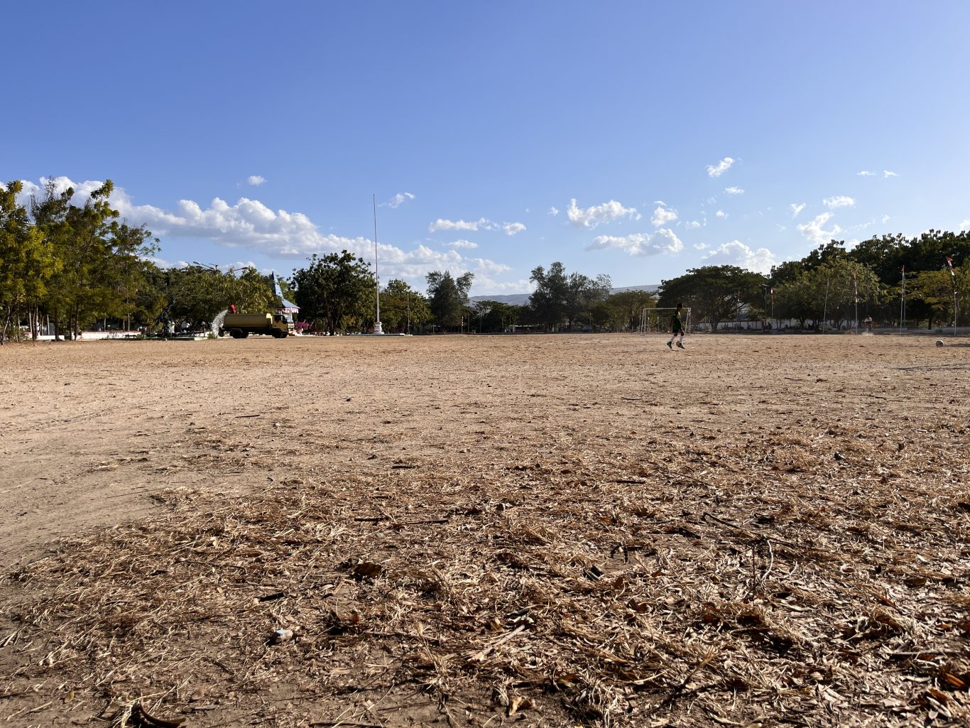 A large, dry, brown field at Heroes Square in Waingapu features a person in a green shirt near a soccer goal, with trees and a flagpole under a blue sky.