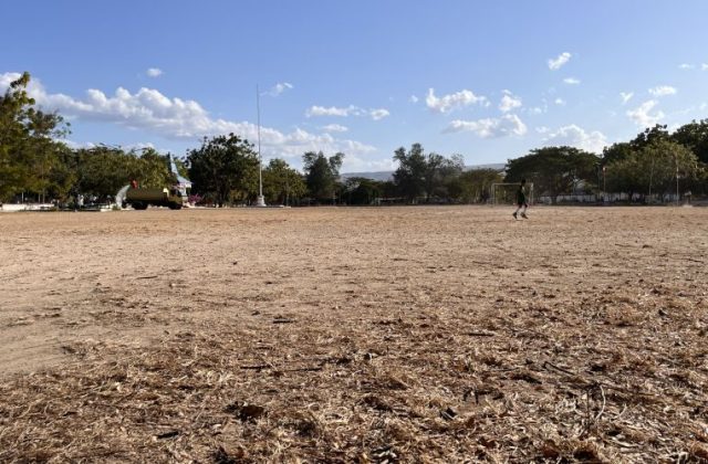 A large, dry, brown field at Heroes Square in Waingapu features a person in a green shirt near a soccer goal, with trees and a flagpole under a blue sky.