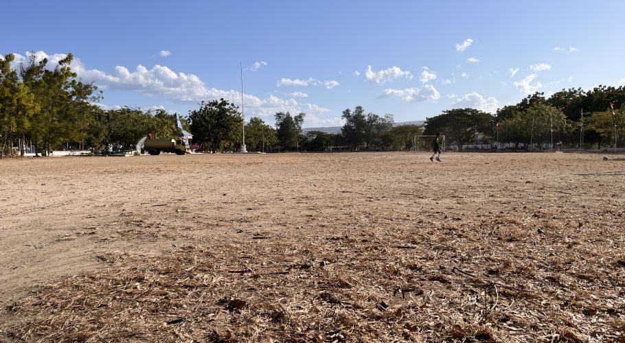 A large, dry, brown field at Heroes Square in Waingapu features a person in a green shirt near a soccer goal, with trees and a flagpole under a blue sky.
