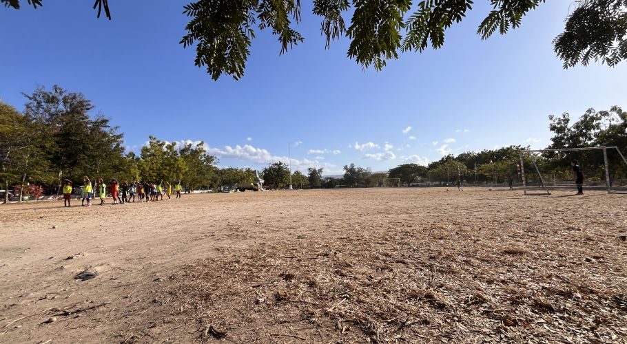 Children in colorful sports gear stretch on a dry, dusty field at Heroes Square in Waingapu under a clear blue sky, with a soccer goal visible.