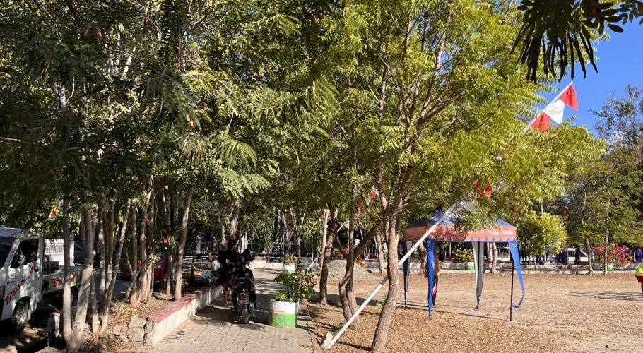 A sunny view of Heroes Square in Waingapu, Sumba, shows green trees, a paved path with vehicles, a blue tent, and red-white flags.