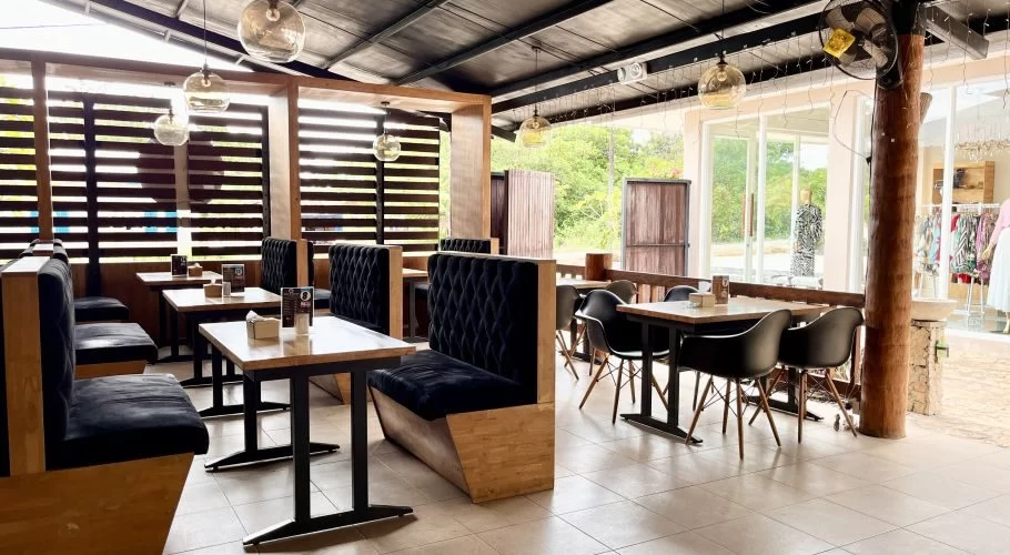 A brightly lit restaurant interior features black upholstered booths, light wooden tables, and black chairs on tiled flooring, with windows looking out to greenery and a shop.