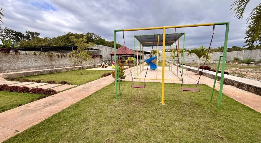 A vibrant children's playground with a green and yellow swing set, a blue slide, and a red-roofed gazebo on a grassy lawn under a cloudy sky.