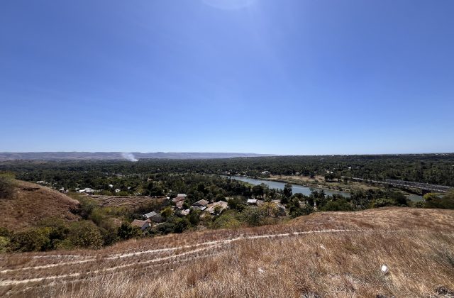 A panoramic view from the dry, grassy Hill of Friendship overlooks Waingapu city, a winding river, and distant hills under a clear blue sky.