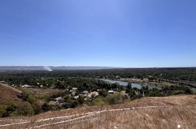 A panoramic view from the dry, grassy Hill of Friendship overlooks Waingapu city, a winding river, and distant hills under a clear blue sky.