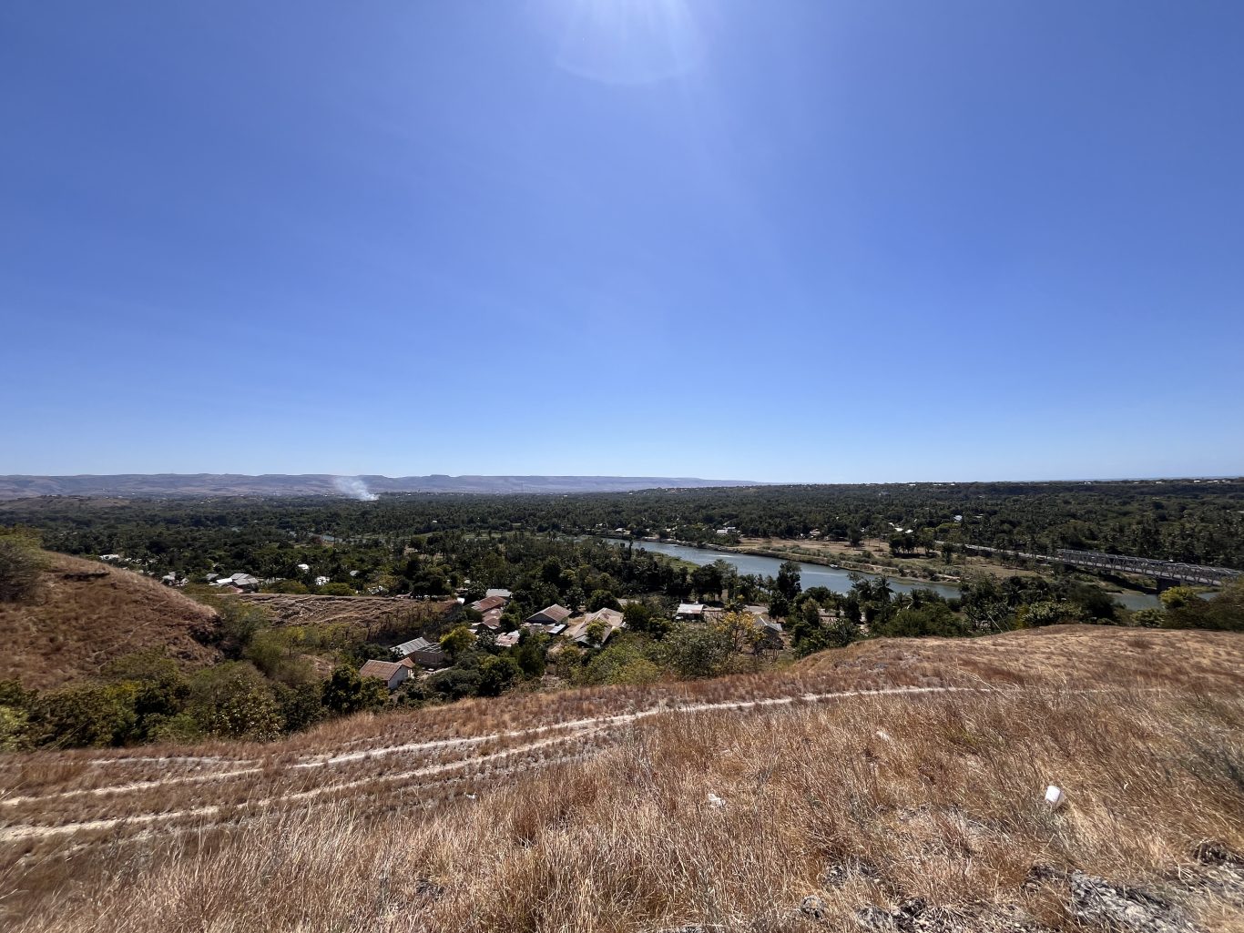 A panoramic view from the dry, grassy Hill of Friendship overlooks Waingapu city, a winding river, and distant hills under a clear blue sky.