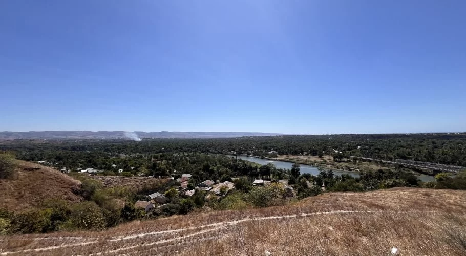 A panoramic view from the dry, grassy Hill of Friendship overlooks Waingapu city, a winding river, and distant hills under a clear blue sky.