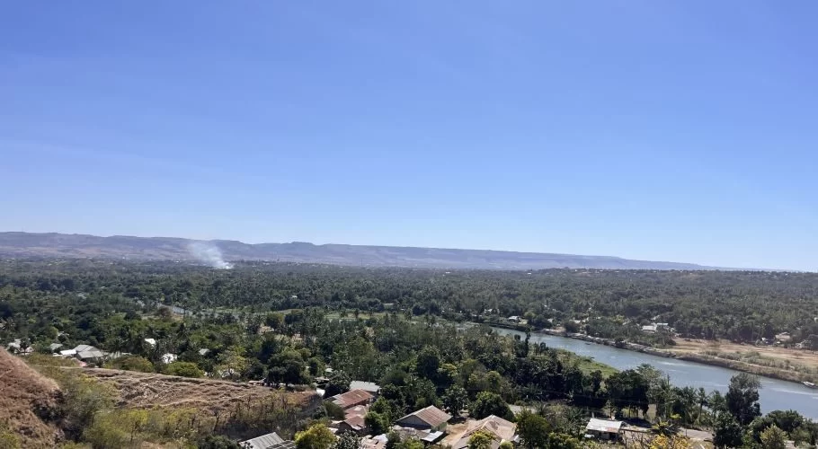 A panoramic view from a dry hillside overlooks a lush green valley with a winding river, scattered houses, distant mountains, and a plume of white smoke under a clear blue sky.