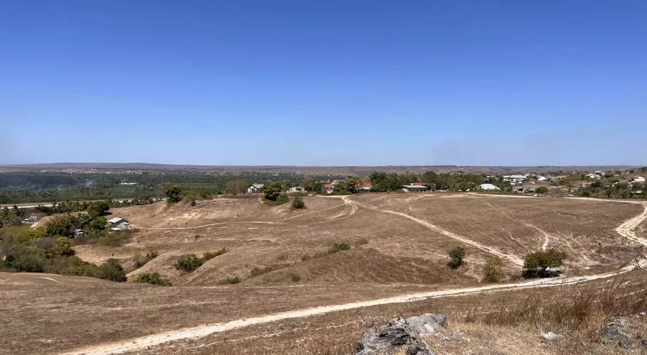A wide panoramic view from a dry, brown hill reveals scattered houses, green trees, and a distant city under a clear blue sky.