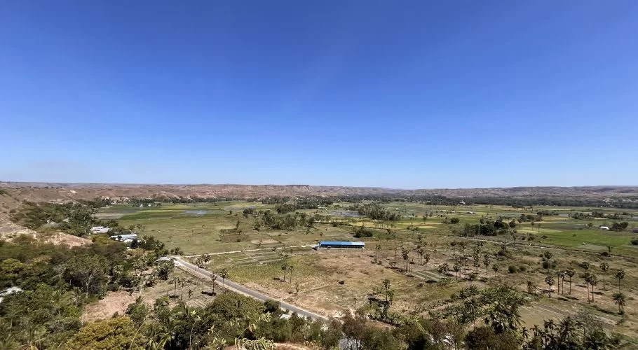 A panoramic view from a hill shows a green valley with scattered trees, a winding road, and distant brown hills under a clear blue sky.