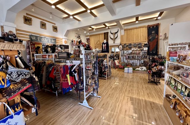 A brightly lit souvenir shop in Sumba, Indonesia, displays colorful clothing with Ikat motifs, bags, jewelry, and wall hangings on racks and shelves.