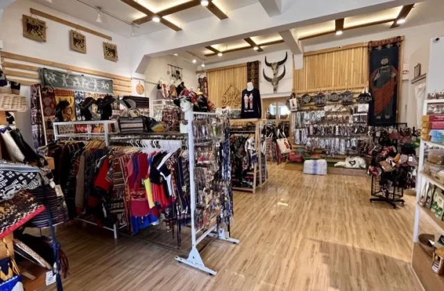 A brightly lit souvenir shop in Sumba, Indonesia, displays colorful clothing with Ikat motifs, bags, jewelry, and wall hangings on racks and shelves.