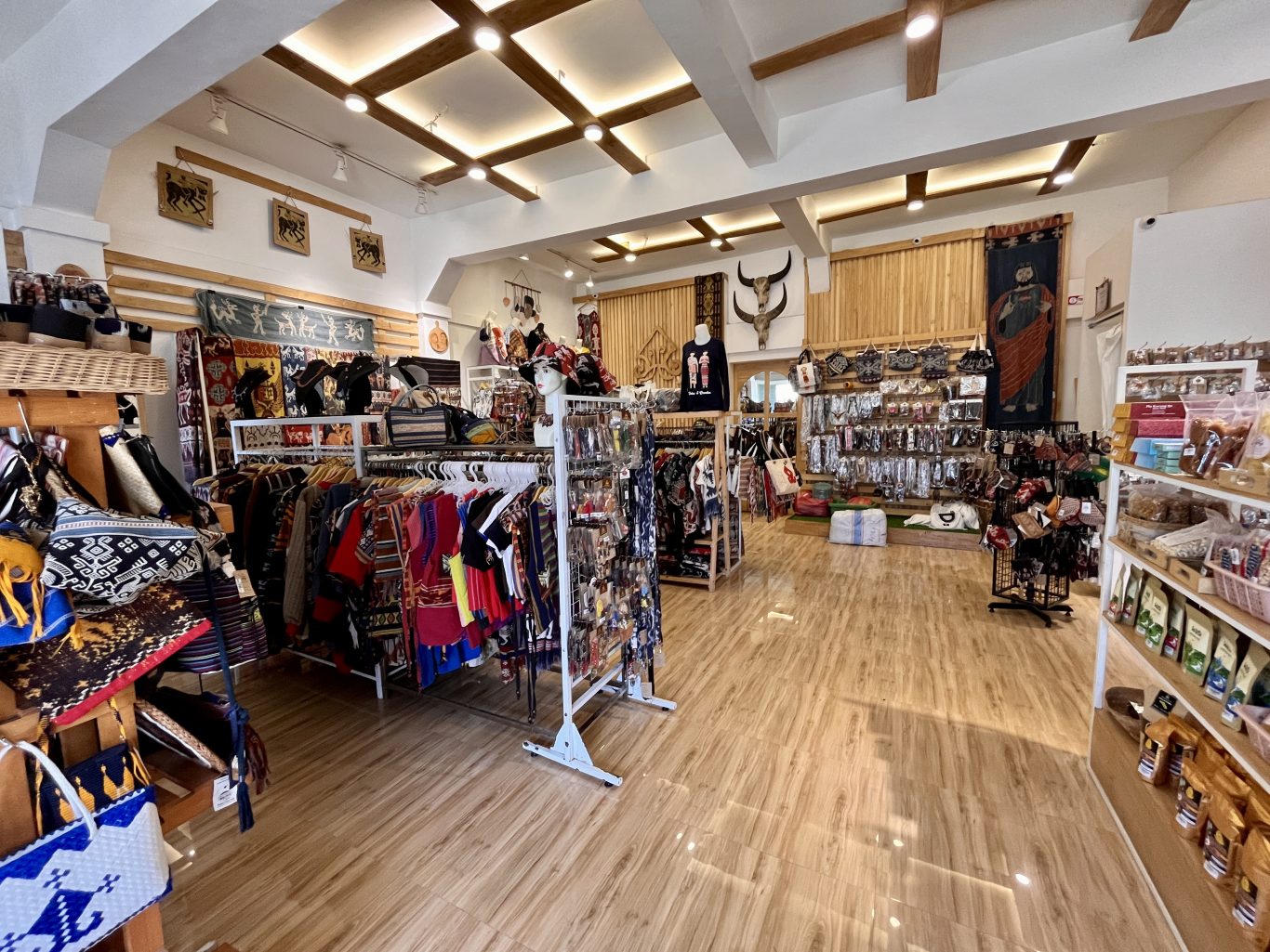 A brightly lit souvenir shop in Sumba, Indonesia, displays colorful clothing with Ikat motifs, bags, jewelry, and wall hangings on racks and shelves.