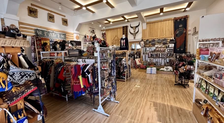 A brightly lit souvenir shop in Sumba, Indonesia, displays colorful clothing with Ikat motifs, bags, jewelry, and wall hangings on racks and shelves.