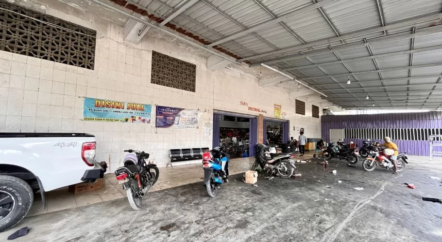 A spacious, open-fronted motorcycle repair shop, Imanuel Motor, in Waikabubak, features several motorcycles being serviced by mechanics, with a white pickup truck parked nearby.