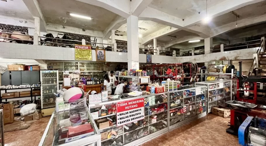 The interior of Imanuel Motor, a spacious motorcycle repair shop in Waikabubak, displays numerous parts and accessories on shelves and in glass cases, with two staff members behind a counter.