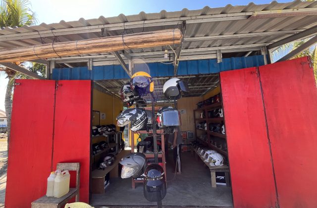 A vibrant red storefront of a helmet shop in Sumba, with open doors revealing numerous colorful helmets displayed on racks inside under a corrugated metal roof.