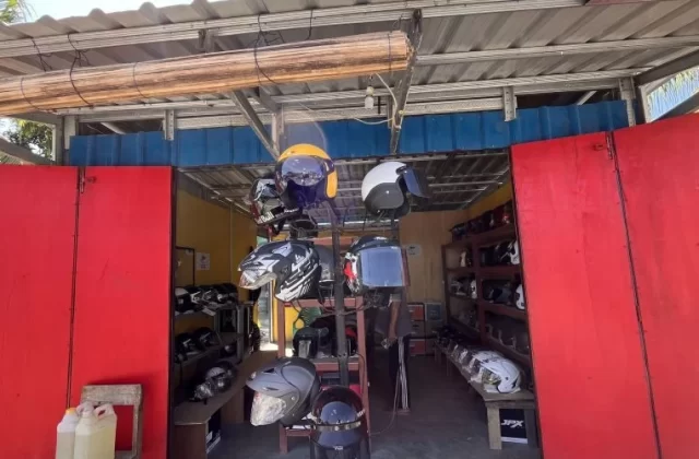 A vibrant red storefront of a helmet shop in Sumba, with open doors revealing numerous colorful helmets displayed on racks inside under a corrugated metal roof.