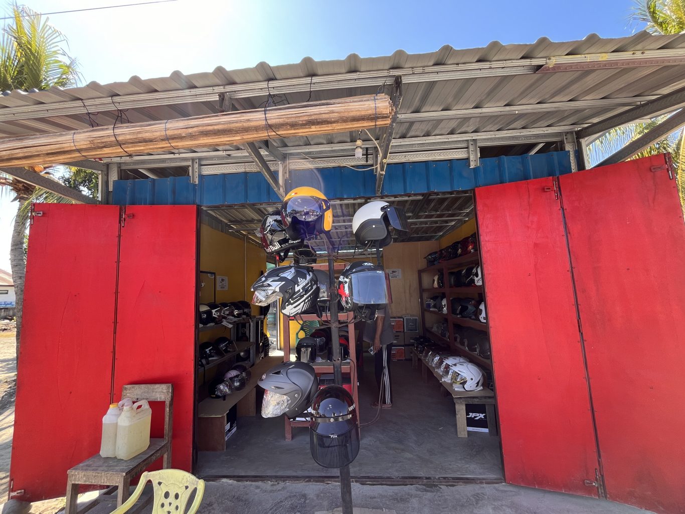 A vibrant red storefront of a helmet shop in Sumba, with open doors revealing numerous colorful helmets displayed on racks inside under a corrugated metal roof.