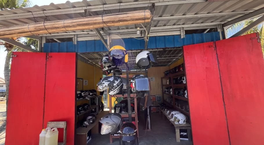 A vibrant red storefront of a helmet shop in Sumba, with open doors revealing numerous colorful helmets displayed on racks inside under a corrugated metal roof.