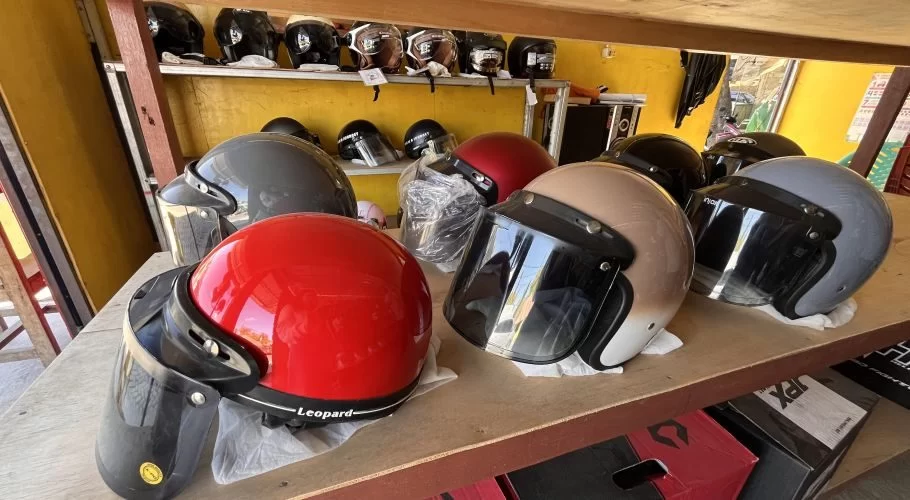 Various motorcycle helmets in red, grey, beige, and black are displayed on wooden shelves inside a store with a bright yellow wall.