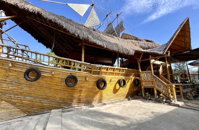 A unique cafe building resembling a wooden ship with a thatched roof and bamboo walls, featuring old tires hanging on its side under a bright blue sky.