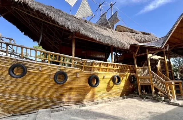 A unique cafe building resembling a wooden ship with a thatched roof and bamboo walls, featuring old tires hanging on its side under a bright blue sky.