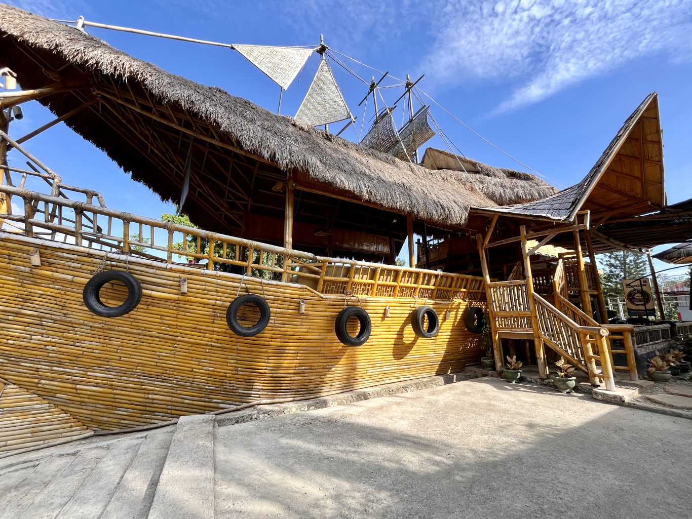 A unique cafe building resembling a wooden ship with a thatched roof and bamboo walls, featuring old tires hanging on its side under a bright blue sky.