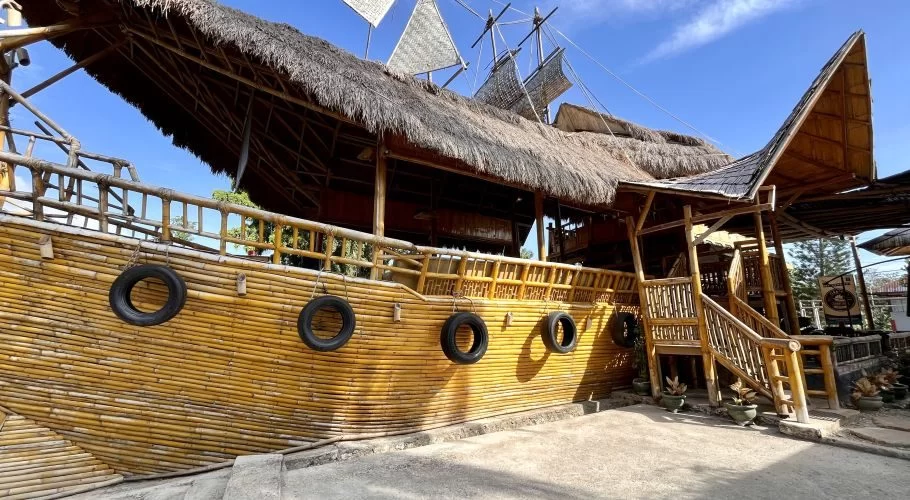 A unique cafe building resembling a wooden ship with a thatched roof and bamboo walls, featuring old tires hanging on its side under a bright blue sky.