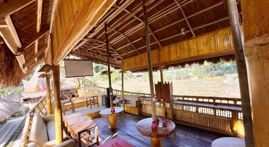 Interior of a cafe, constructed from light brown bamboo and wood, featuring a thatched roof, various seating, and a projector screen, overlooking an open field.