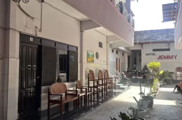 The exterior of Jemmy Hotel in Waingapu features a light-colored two-story building with dark doors, wooden chairs, and potted plants along a sunny walkway.