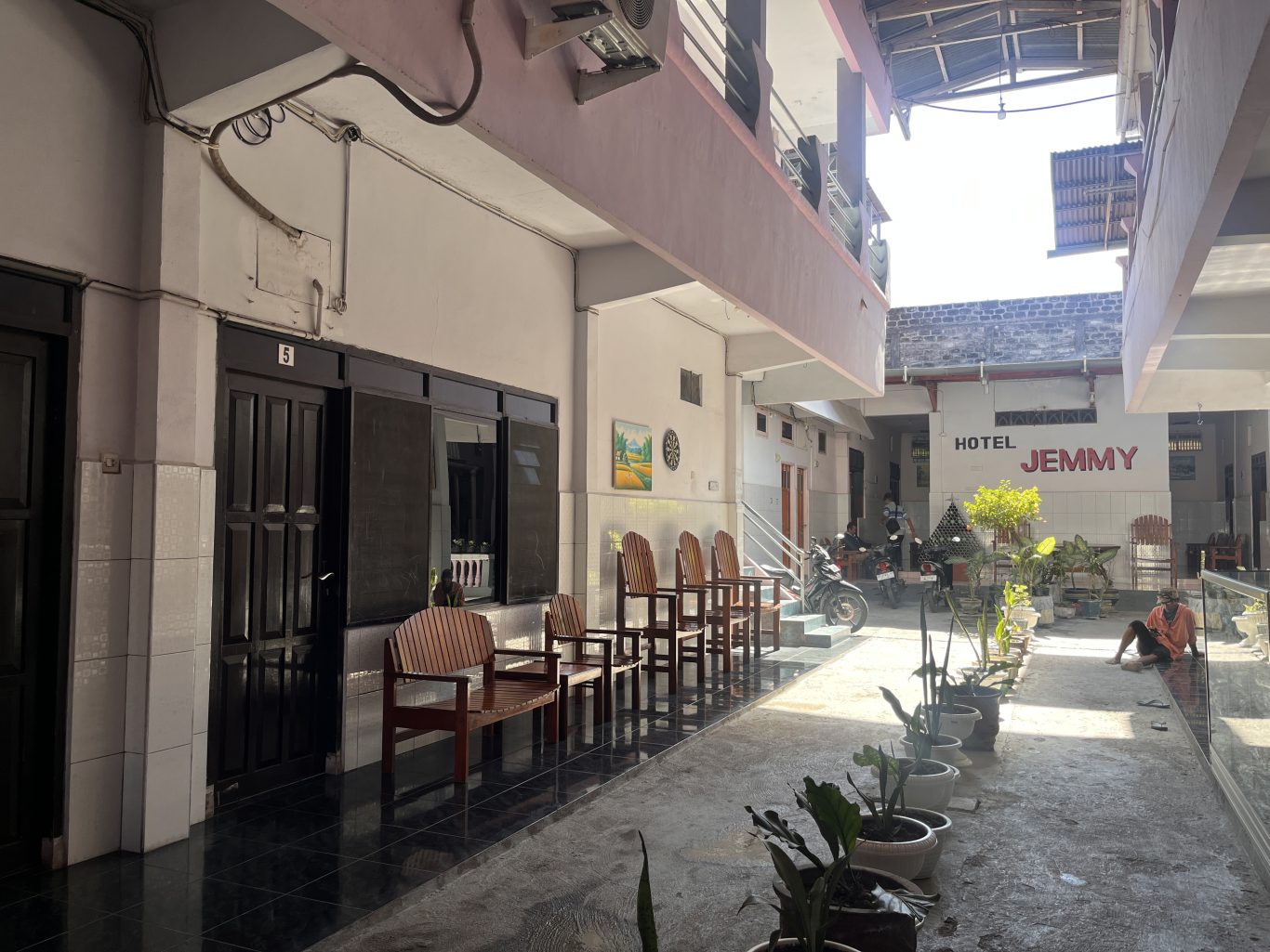 The exterior of Jemmy Hotel in Waingapu features a light-colored two-story building with dark doors, wooden chairs, and potted plants along a sunny walkway.