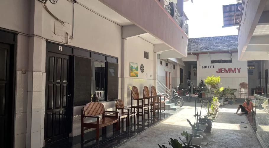 The exterior of Jemmy Hotel in Waingapu features a light-colored two-story building with dark doors, wooden chairs, and potted plants along a sunny walkway.