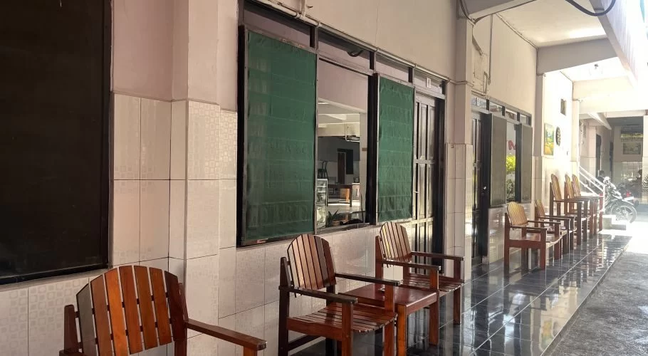 A long, sunlit outdoor hallway with several wooden chairs and small tables lined against a tiled wall with green-shuttered windows.
