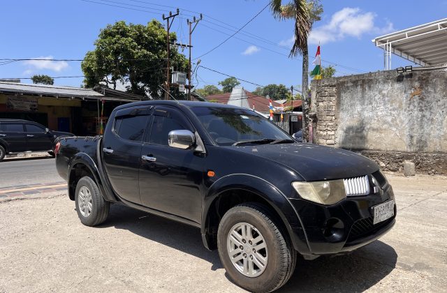 A black Mitsubishi Triton pickup truck is parked on a dirt shoulder next to a paved road in a sunny town setting, with buildings and trees in the background.