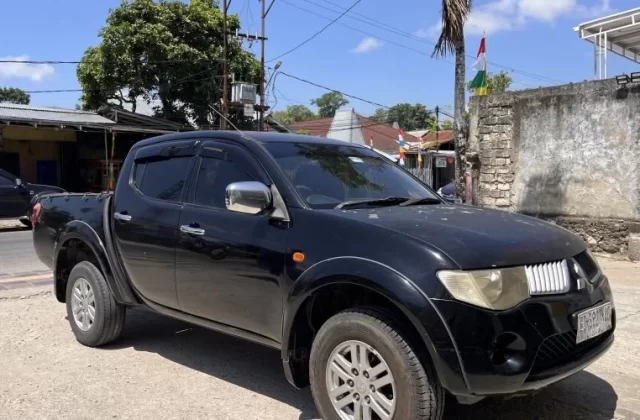 A black Mitsubishi Triton pickup truck is parked on a dirt shoulder next to a paved road in a sunny town setting, with buildings and trees in the background.