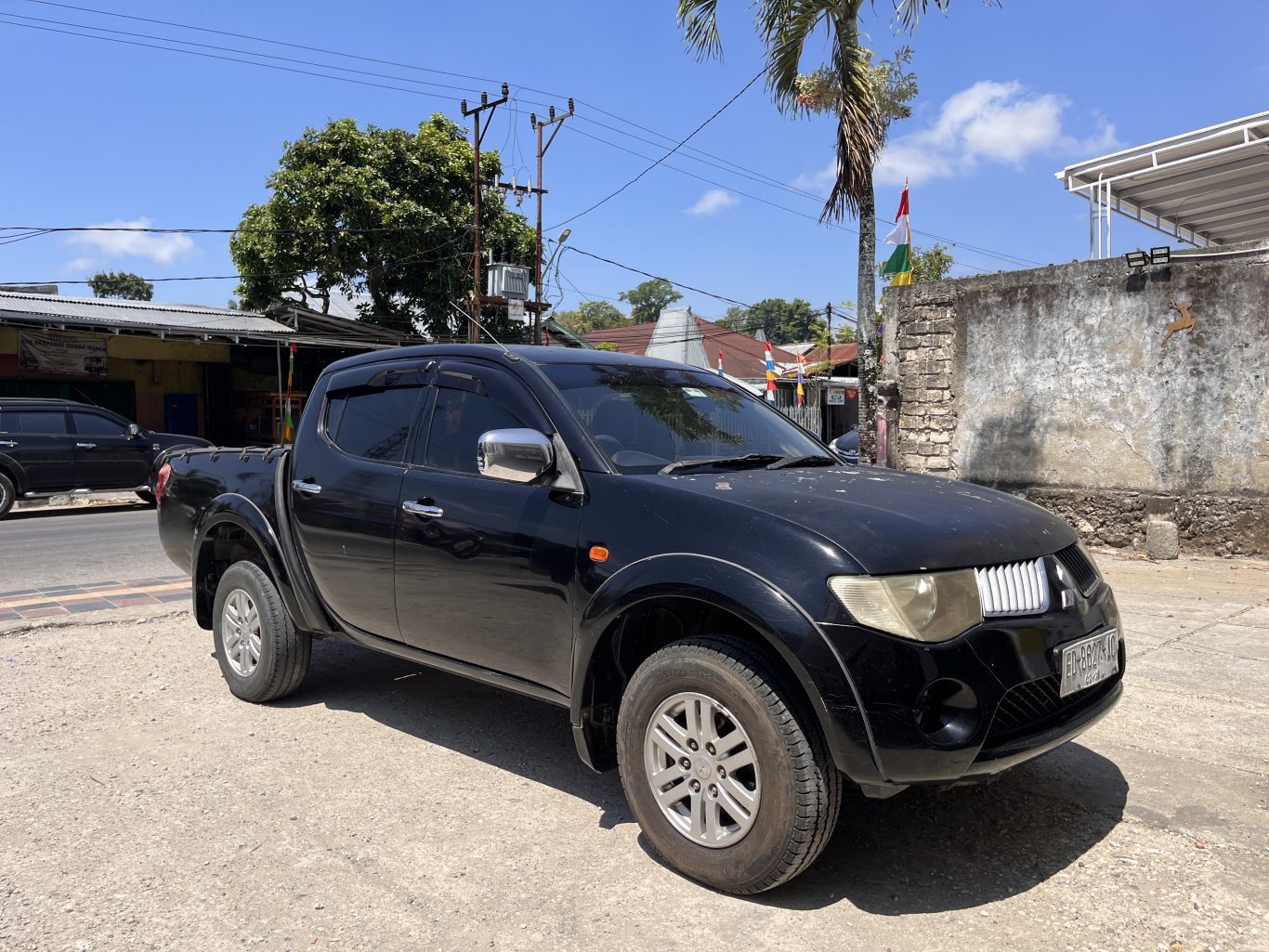 A black Mitsubishi Triton pickup truck is parked on a dirt shoulder next to a paved road in a sunny town setting, with buildings and trees in the background.