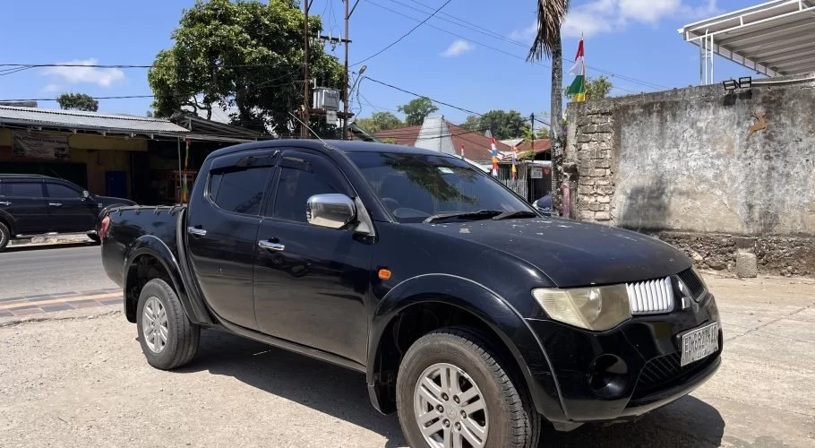 A black Mitsubishi Triton pickup truck is parked on a dirt shoulder next to a paved road in a sunny town setting, with buildings and trees in the background.