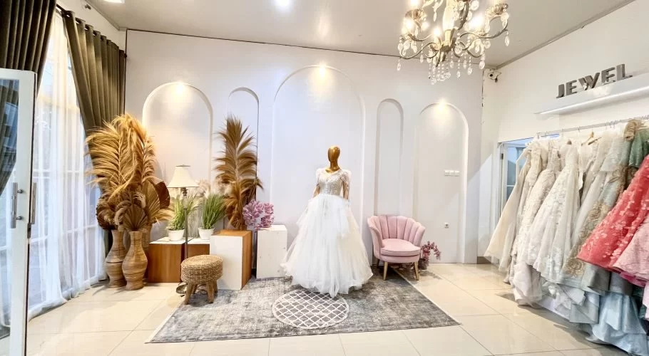 A bright bridal shop interior features a mannequin in a white wedding dress, surrounded by decorative dried palms, a pink armchair, and racks of colorful gowns.