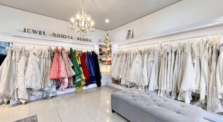 Brightly lit bridal shop interior featuring racks of white and colorful wedding and formal dresses, with a tufted bench and chandelier.